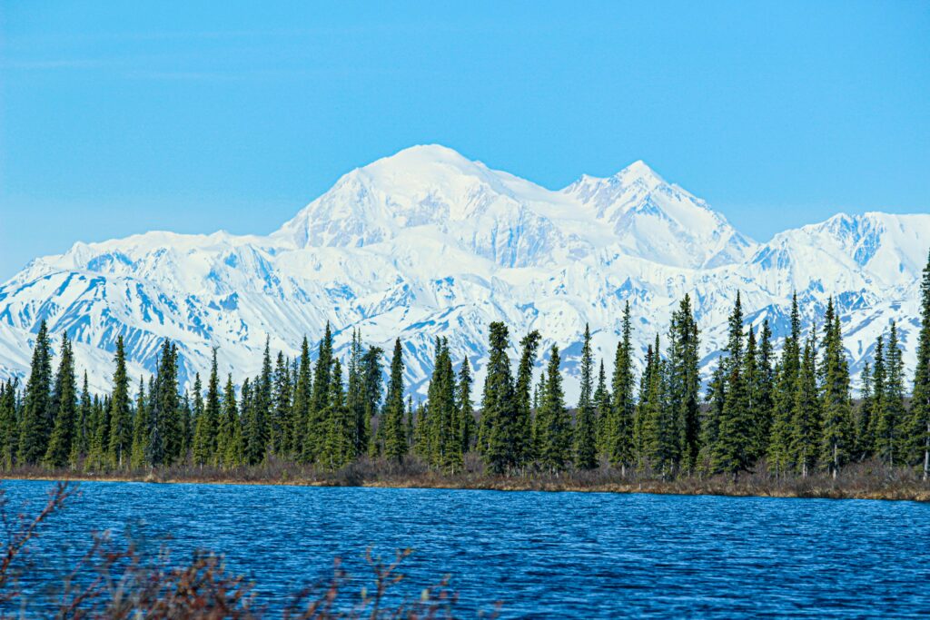 A scenic view of a blue lake and a line of spruce trees against the backdrop of massive snow-covered peaks in the Prince of Wales Island wilderness.